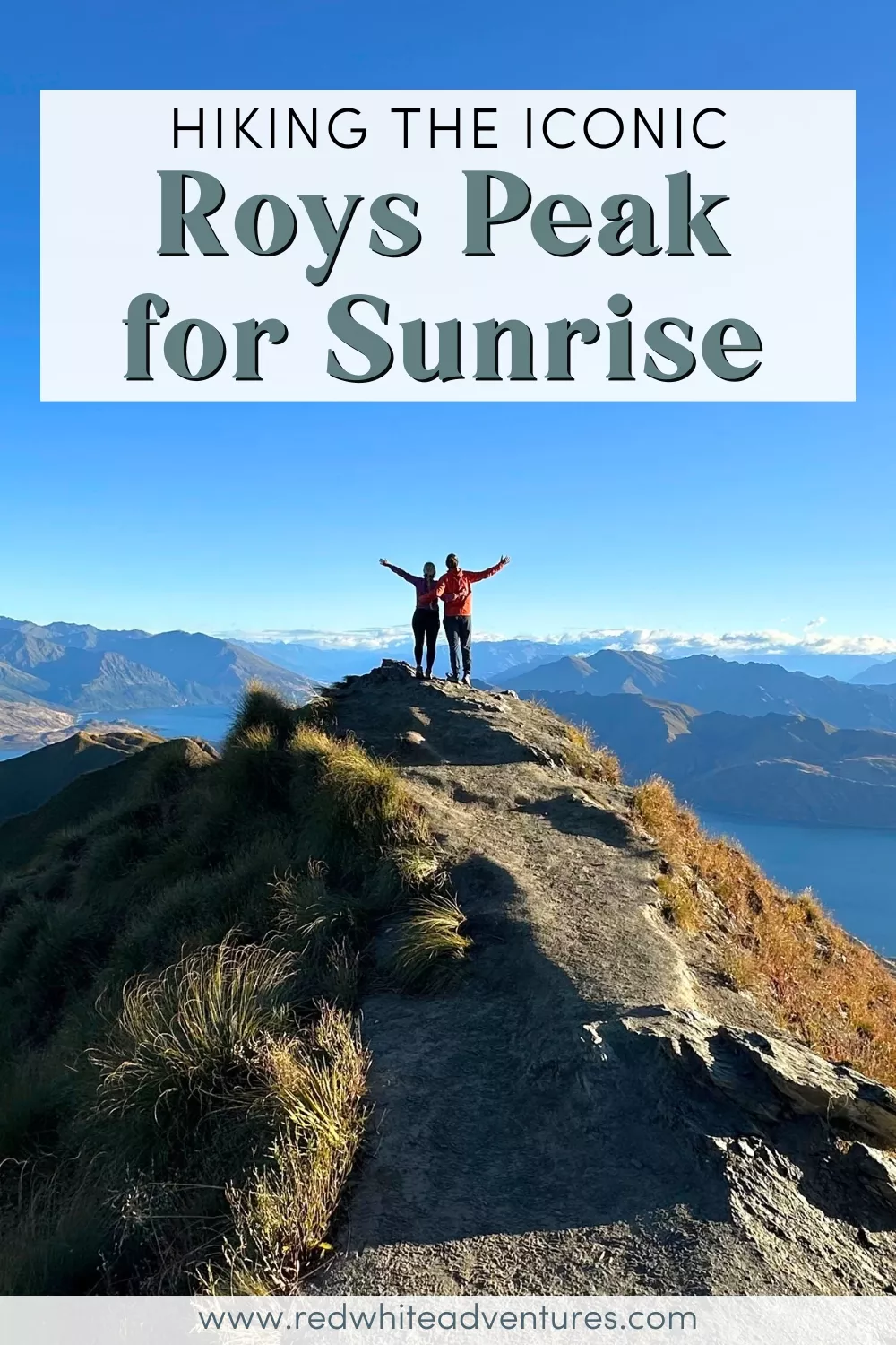 Two hikers stand triumphantly with arms raised on a narrow ridge at Roys Peak, overlooking a breathtaking view of Lake Wānaka and surrounding mountains under a clear blue sky. Text above reads “Hiking the iconic Roys Peak for Sunrise”.