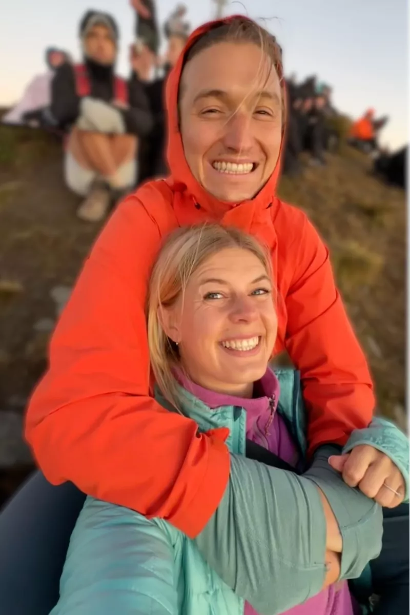 A smiling couple bundled in colourful jackets takes a close-up selfie at sunrise on Roys Peak, with one hugging the other from behind. In the background, a group of hikers sits along the ridgeline, waiting for the sun to rise.