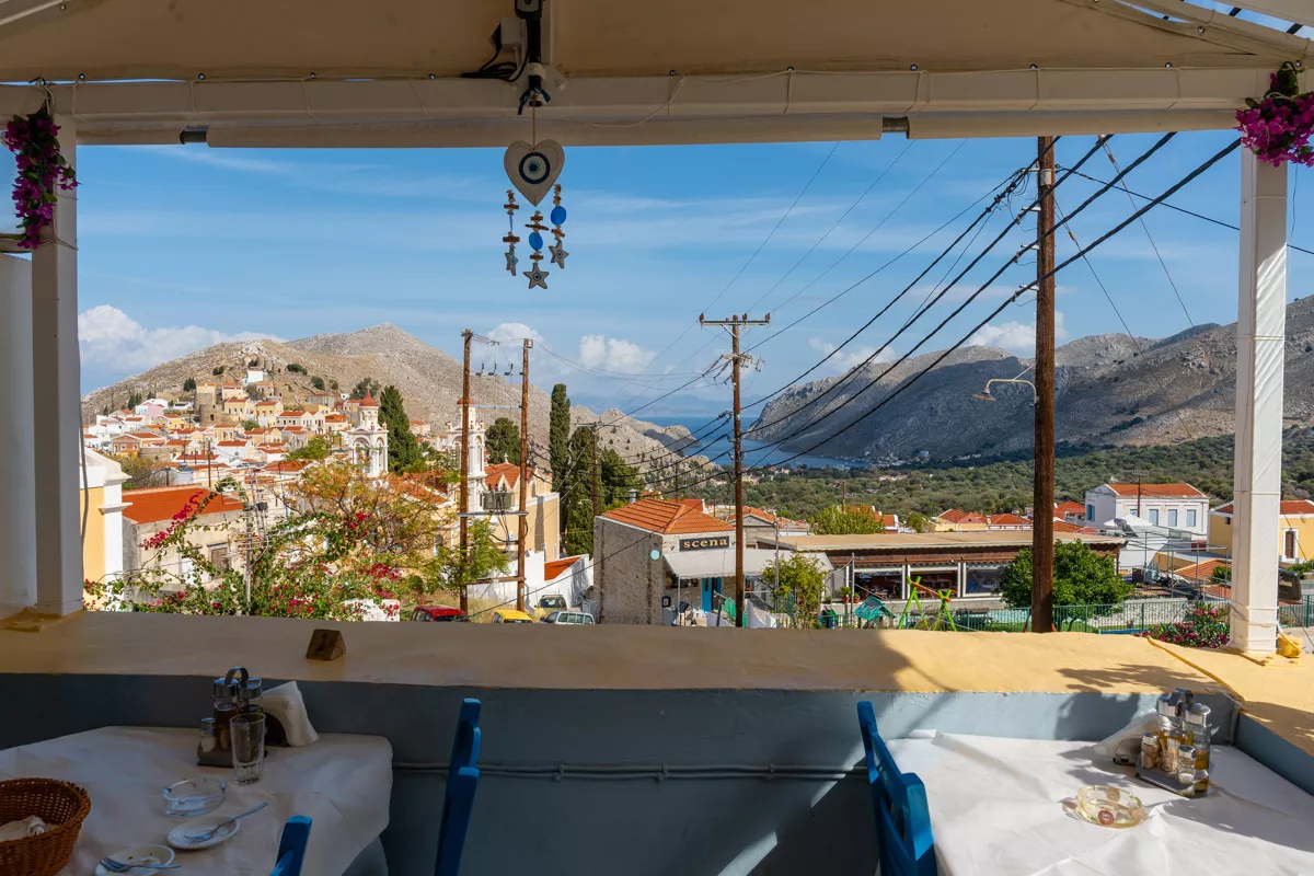 View from Taverna Zoe in Symi, Greece, looking out over red-roofed homes, church towers, and distant mountains with a glimpse of the sea. The photo is framed by hanging decor and set tables, capturing the relaxed charm of this hillside restaurant.