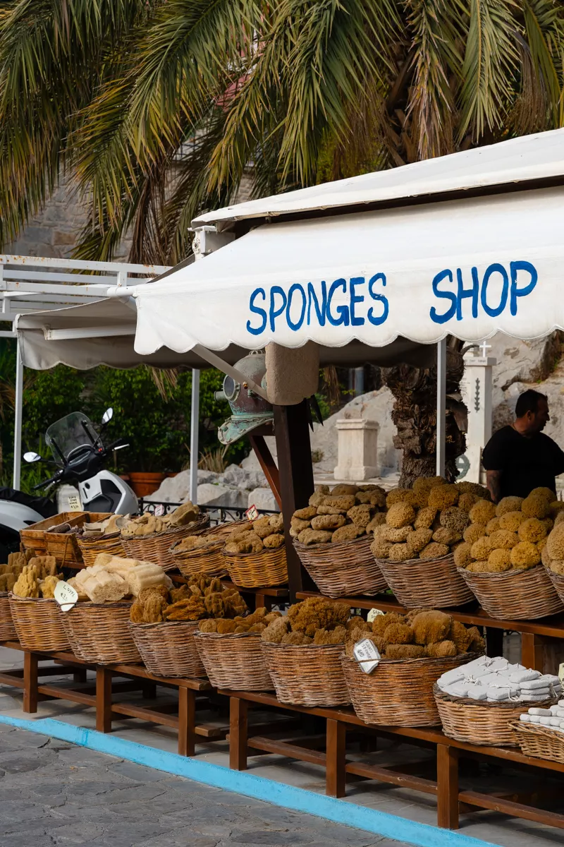A traditional sponge shop in Symi displays baskets overflowing with natural sea sponges under a white canopy sign. Palm trees and a vintage diving helmet add to the island charm.