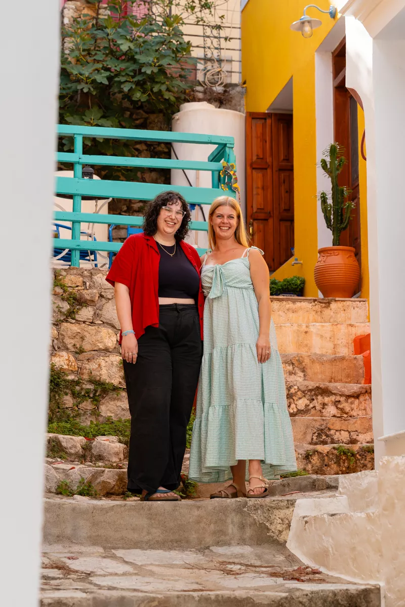 Two smiling women pose in a colourful alleyway in Symi, surrounded by bright houses, a turquoise railing, and a terracotta pot with a cactus. The atmosphere is warm, cheerful, and welcoming.