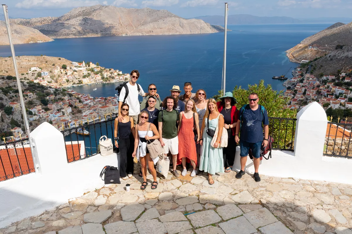 A group of smiling travellers poses at a scenic lookout above Symi, with sweeping views of the harbour, pastel houses cascading down the hillside, and the deep blue sea stretching to rugged mountains in the distance.