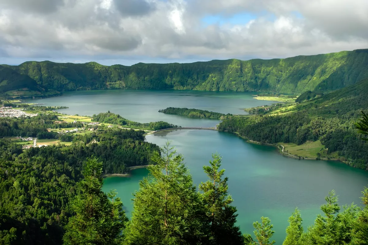 A panoramic view of Sete Cidades, a twin-lake volcanic crater on São Miguel Island in the Azores, with lush green hills and a small village along the lakeshore under a moody sky.