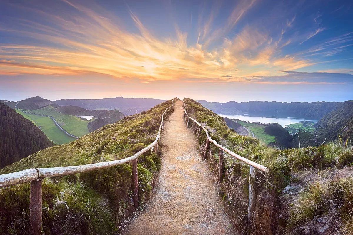 A hikinh trail with rustic wooden railings leads along a ridgeline at sunset, overlooking the Sete Cidades crater lakes on São Miguel Island in the Azores. The glowing sky and sweeping coastal views highlight one of the most iconic hiking spots in the archipelago.