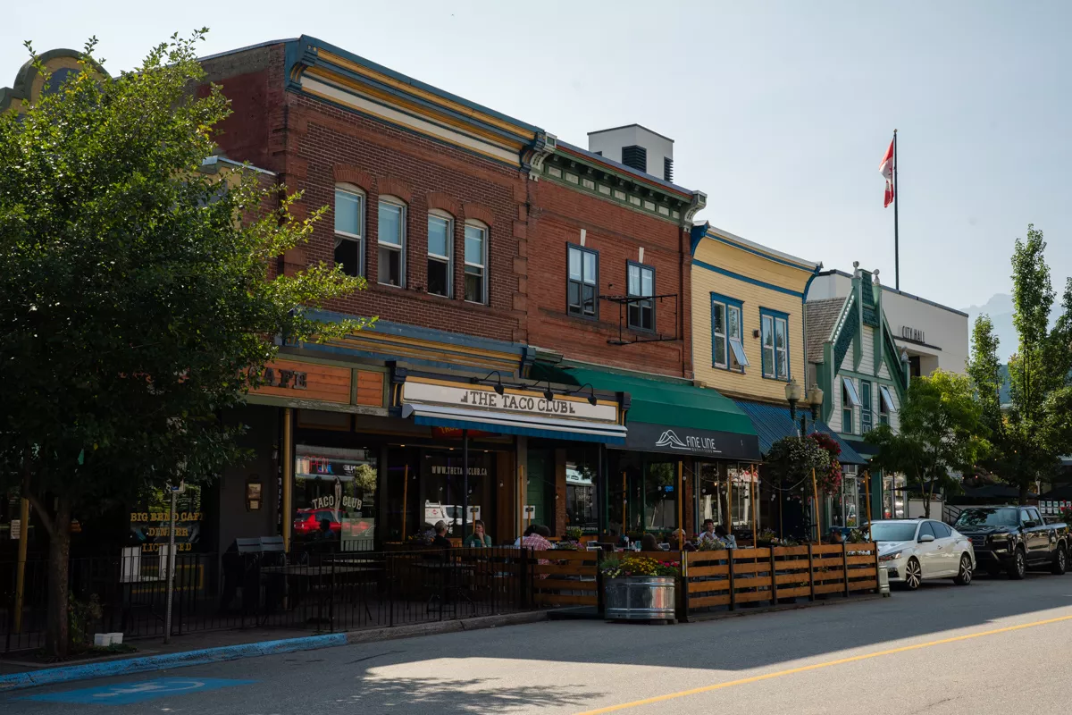 A lively downtown street in Revelstoke featuring colourful historic buildings, including The Taco Club restaurant with outdoor seating and a Canadian flag flying above the city hall. Diners enjoy a sunny day in this charming small town with a backdrop of distant mountains.
