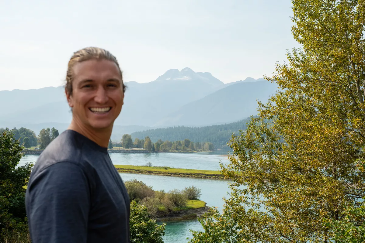 A smiling man stands in front of a peaceful river landscape with forested banks and a small island, backed by the towering silhouette of Mt Begbie. The mountain’s rugged peaks are partially covered with glaciers, adding a dramatic alpine touch to the scenic view.