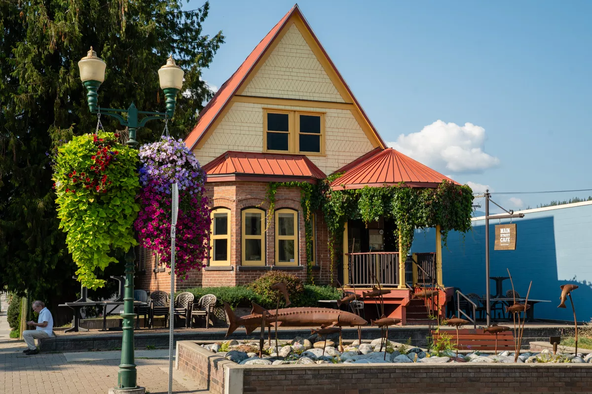 A charming café in a historic A-frame house with a red metal roof and vine-covered porch, framed by hanging flower baskets and a small outdoor seating area. A whimsical metal fish sculpture garden decorates the front, adding character to this inviting spot named Main Street Café.