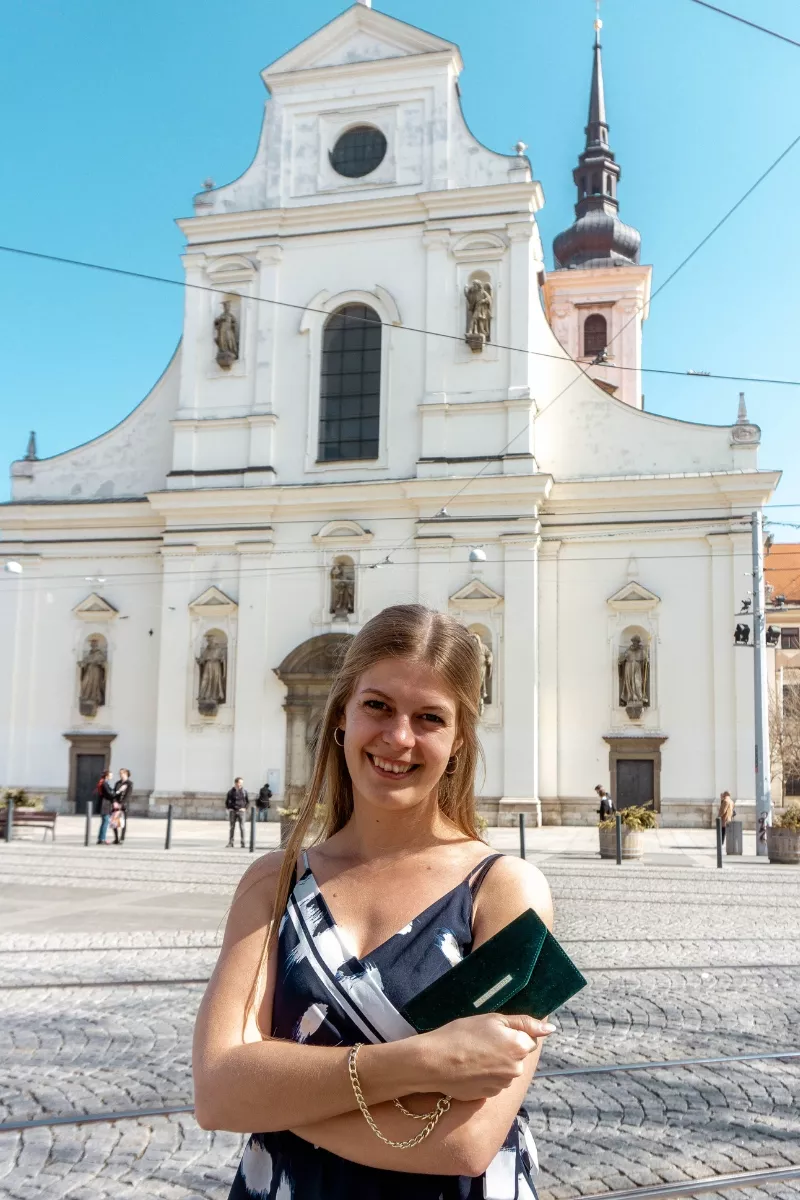 A woman smiling and standing in front of a large white historic church with statues on the facade and a tall tower in the background.