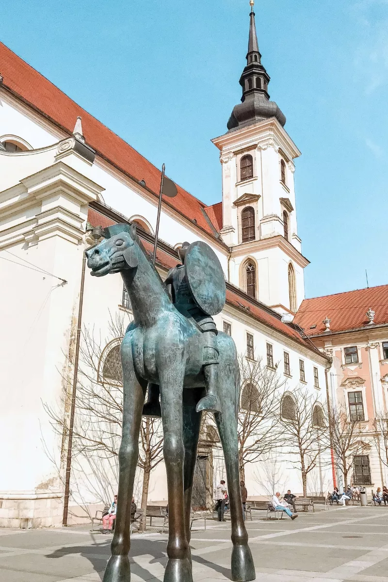 A tall, thin horse statue with a rider holding a shield in front of a white church with a red roof and a tower, located in a sunny square.