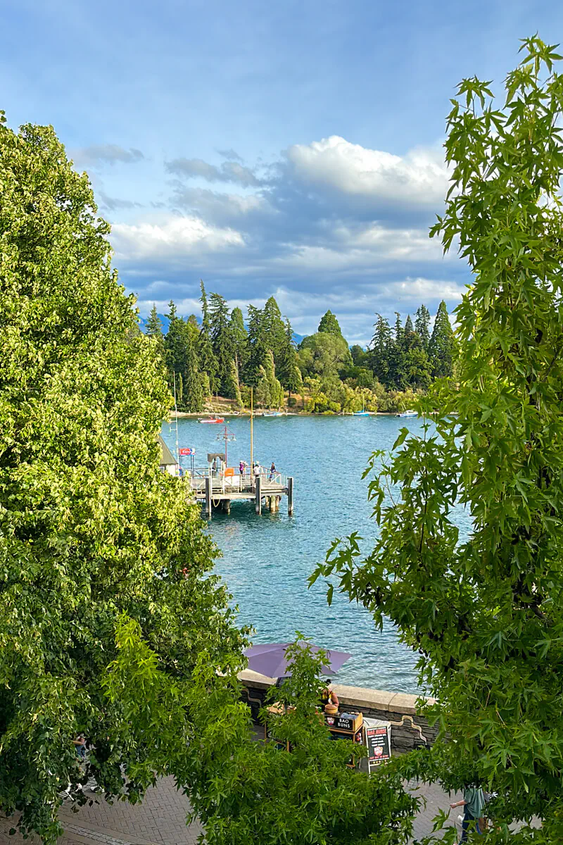 A view of the Queenstown waterfront on Lake Wakatipu, framed by leafy green trees. A wooden jetty with people standing on it extends into the turquoise lake, with a forested shoreline and mountains in the background under a partly cloudy sky.