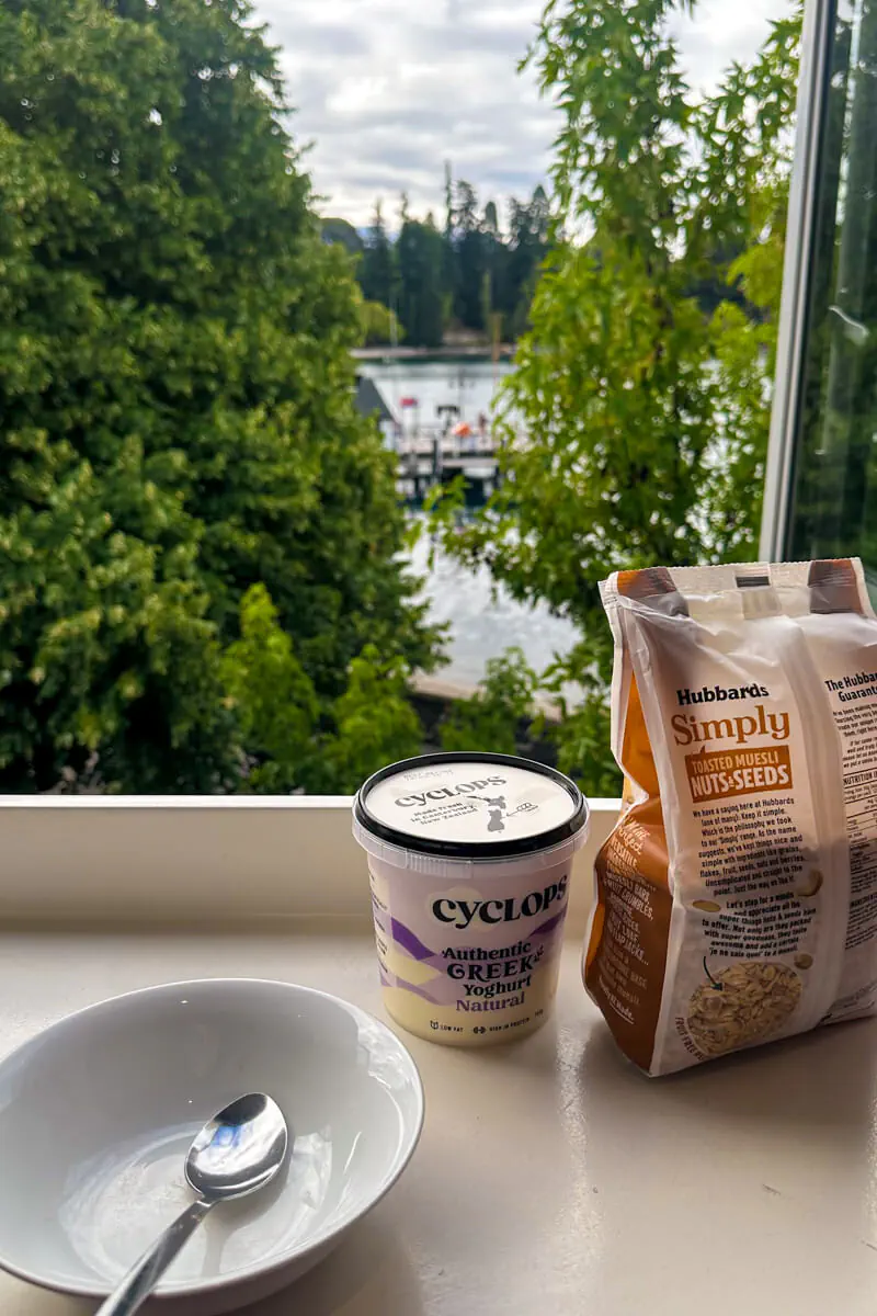 A container of Cyclops Greek yoghurt, a bag of Hubbard’s Simply Toasted Muesli, and a white bowl with a spoon sit on a windowsill. Through the window, lush green trees and part of a dock on Lake Wakatipu in Queenstown are visible.