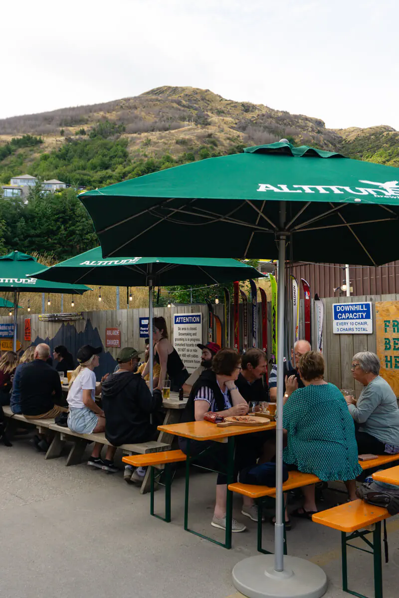 An outdoor seating area at Altitude Brewing, with people gathered at picnic tables under green umbrellas branded with the brewery’s logo. Behind them, ski gear decorates the fence, and hills rise in the background.