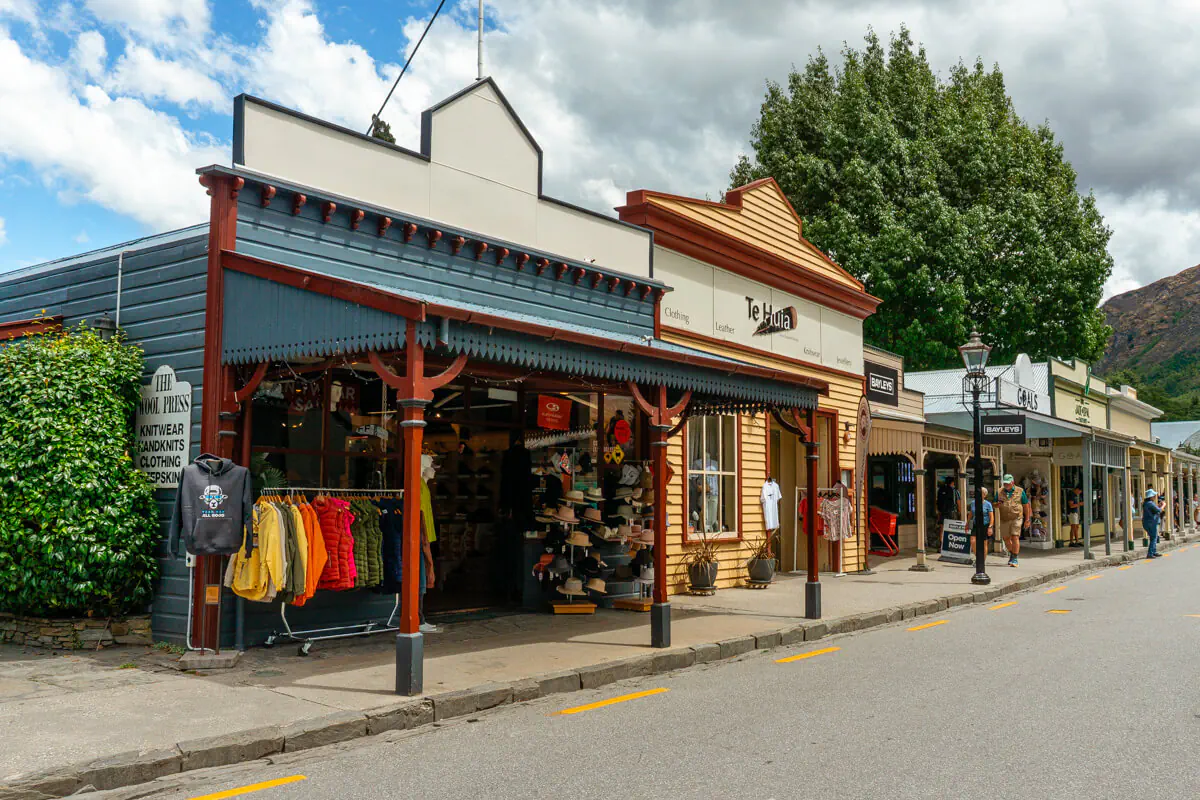 A charming street in Arrowtown, New Zealand, features quaint historic storefronts with colourful signage and wooden facades. Shops like "The Kiwi Press" and "Te Huia" display clothing and hats outside under shaded awnings. A few people stroll along the sidewalk.
