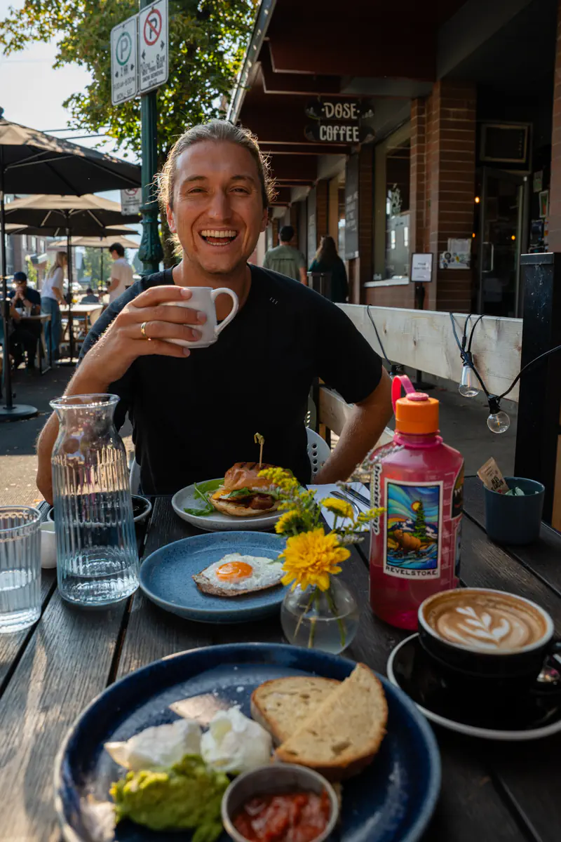 A cheerful outdoor brunch scene at Dose Coffee in Revelstoke, BC. A man is sitting at a wooden patio table, smiling brightly while holding a cup of coffee. In front of him are delicious-looking breakfast dishes – including toast, poached eggs with avocado, a fried egg on crispbread, and a breakfast sandwich. A latte with pretty foam art and a bright pink water bottle add pops of color to the table.