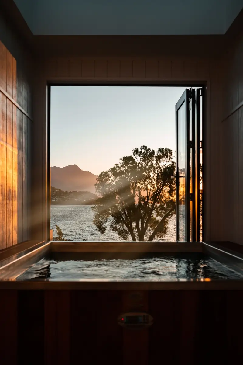 Indoor hot tub with wooden panelling and open glass doors framing a tranquil lakeside view of Lake Wakatipu in Queenstown at sunset. Warm golden light streams through a tree, with distant mountains silhouetted against the sky.