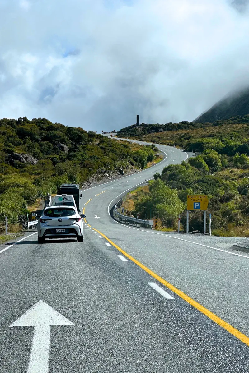 A winding mountain highway leading uphill toward low-lying clouds, with a white car driving ahead on the two-lane road. Green shrubs and rocky slopes line the roadside, and a yellow sign on the right points to the Temple Basin ski area. The scene captures the dramatic alpine drive through New Zealand’s Southern Alps.