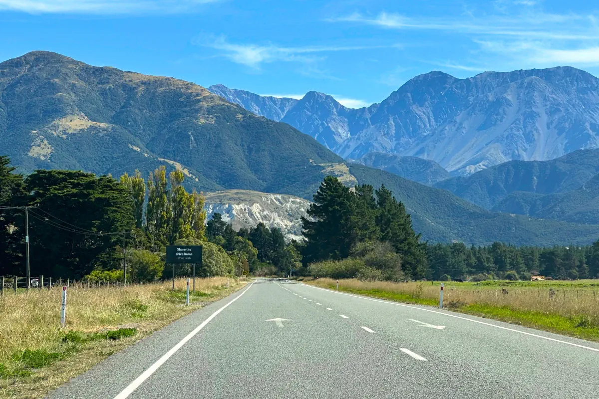 A rural highway stretches toward tall, rugged mountains under a clear blue sky. The road is lined with grass, trees, and a roadside sign that reads “Share the drive home.” The towering peaks of New Zealand’s Southern Alps rise dramatically in the background.