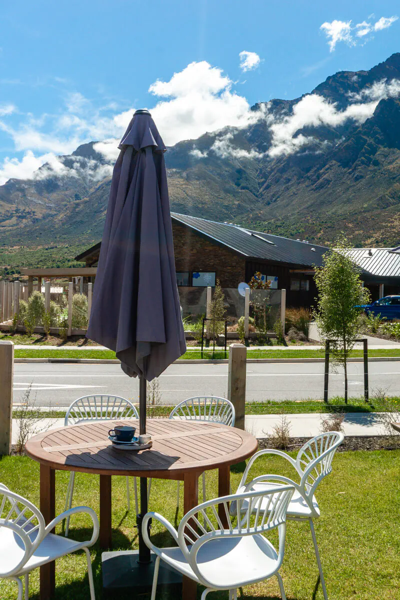 A round outdoor table with white chairs and a closed umbrella on a grassy lawn, set against a backdrop of modern alpine-style buildings and stunning mountains partially covered by clouds in Queenstown.