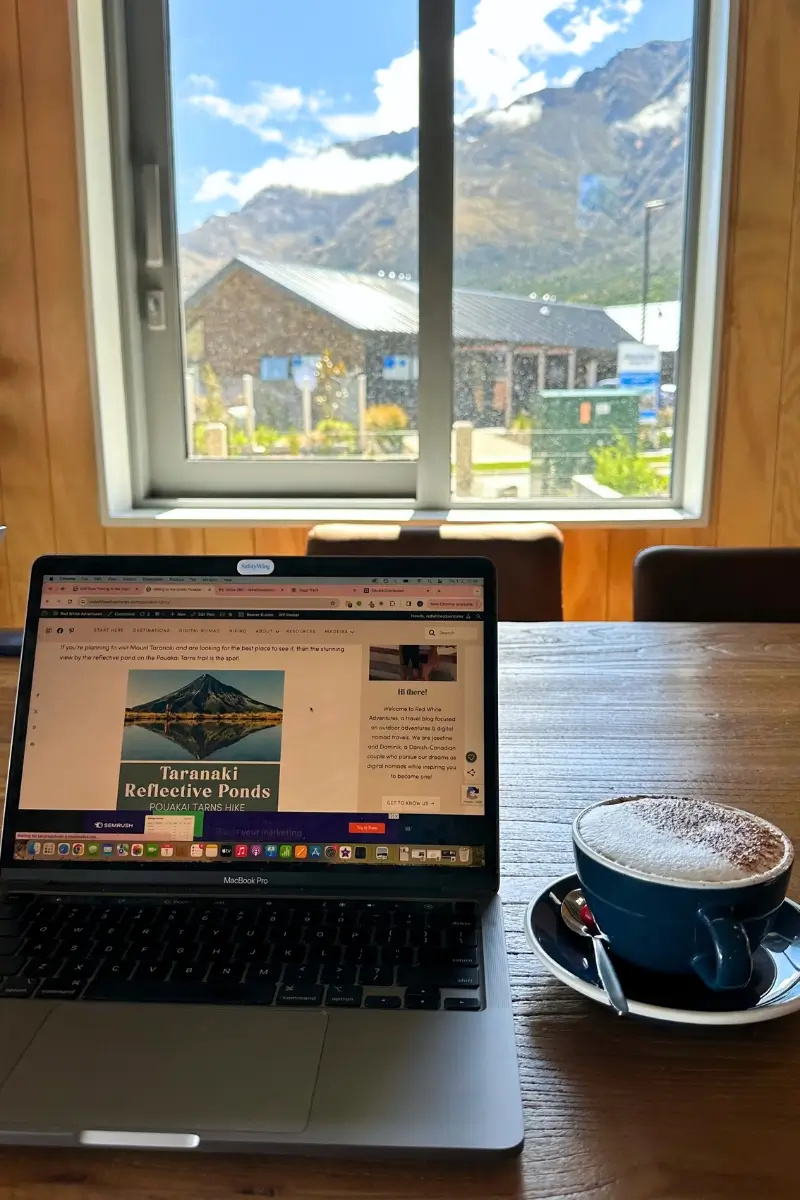 A cozy workspace setup inside a café features an open laptop displaying a travel blog article titled "Taranaki Reflective Ponds." Beside the laptop is a blue cup of cappuccino with cocoa powder on top. Through the window in the background, there’s a sunny view of a rustic building, lush greenery, and towering mountains under a partly cloudy sky.