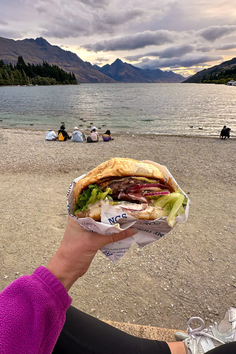 A hand holds up a large, loaded burger wrapped in paper with Lake Wakatipu and the Remarkables mountain range in the background at sunset in Queenstown, New Zealand. Several people sit along the pebbled lakeshore, enjoying the view.