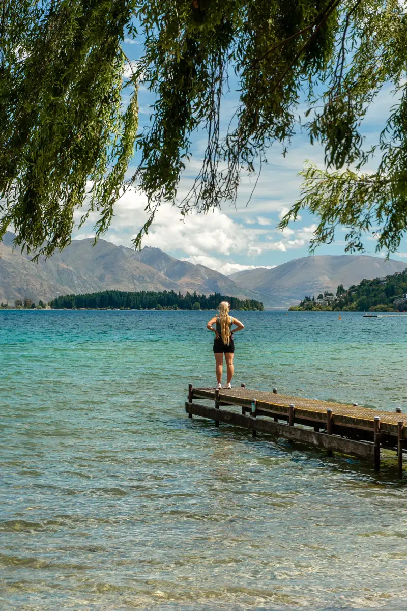 A woman with long blonde hair stands on a wooden dock overlooking the clear turquoise waters of Lake Wakatipu, New Zealand. Mountains rise in the distance under a partly cloudy sky, with leafy tree branches framing the top of the image.