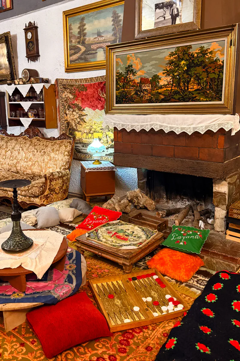 Cosy interior of a traditional Greek café with vintage decor, tapestries, patterned pillows, board games, and a stone fireplace surrounded by antiques and warm lighting.