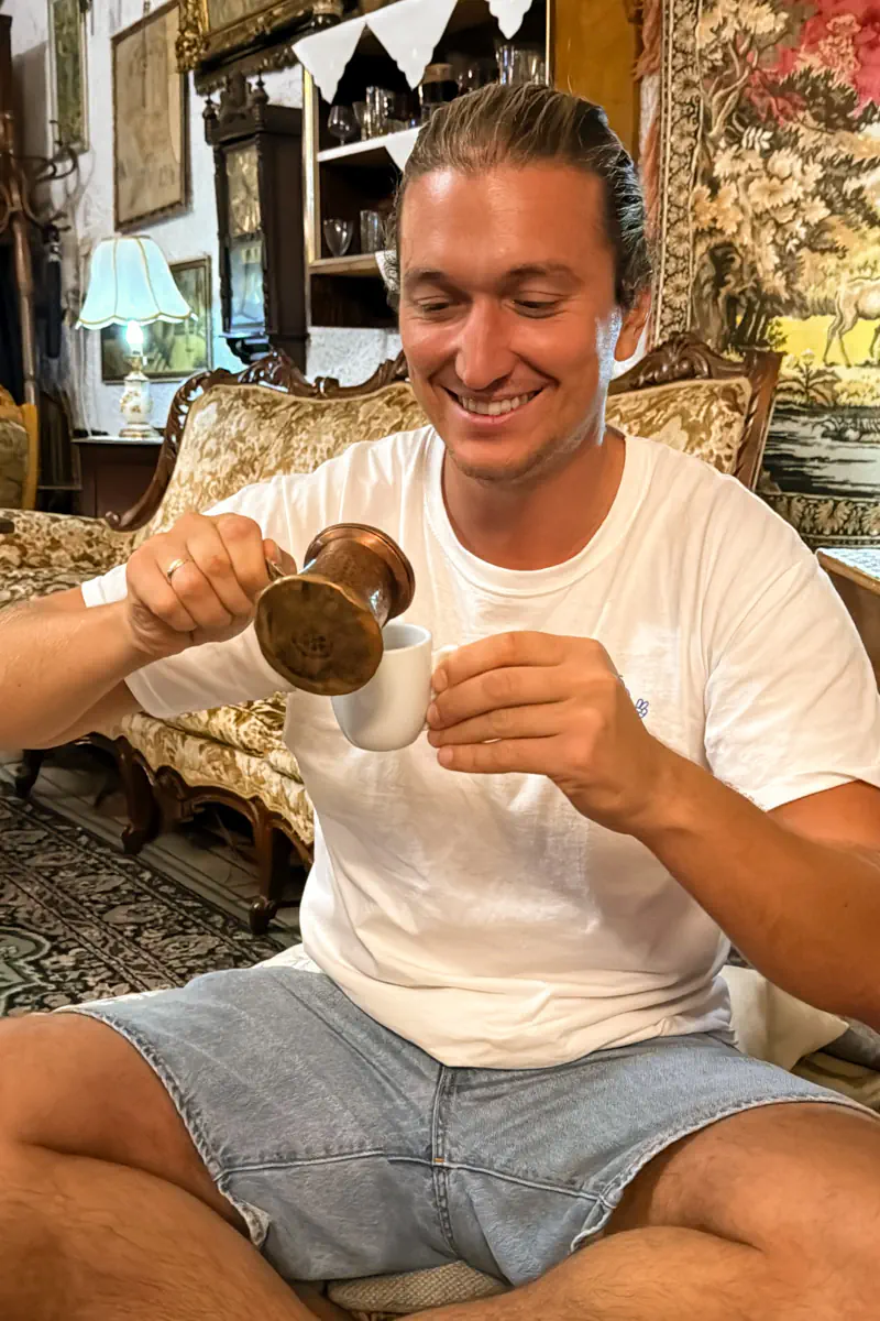 A smiling man in casual clothes pours traditional Greek coffee into a small white cup inside a vintage-style café with ornate furniture and wall tapestries.