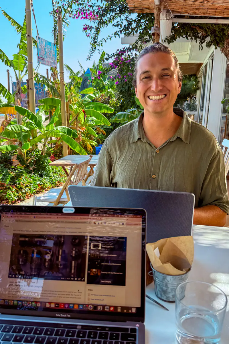 A smiling man works on his laptop at an outdoor café surrounded by banana plants and tropical greenery in Rhodes, Greece. A second laptop in the foreground shows a browser open, with a glass of water on the table.