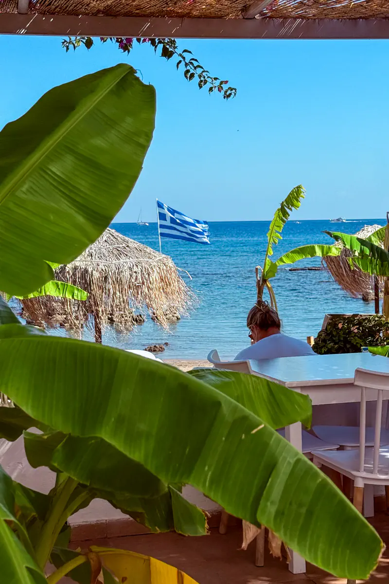 A peaceful beachfront café scene with a person sitting at a white table under thatched umbrellas, framed by large banana leaves. A Greek flag waves above the bright blue Aegean Sea in the background.
