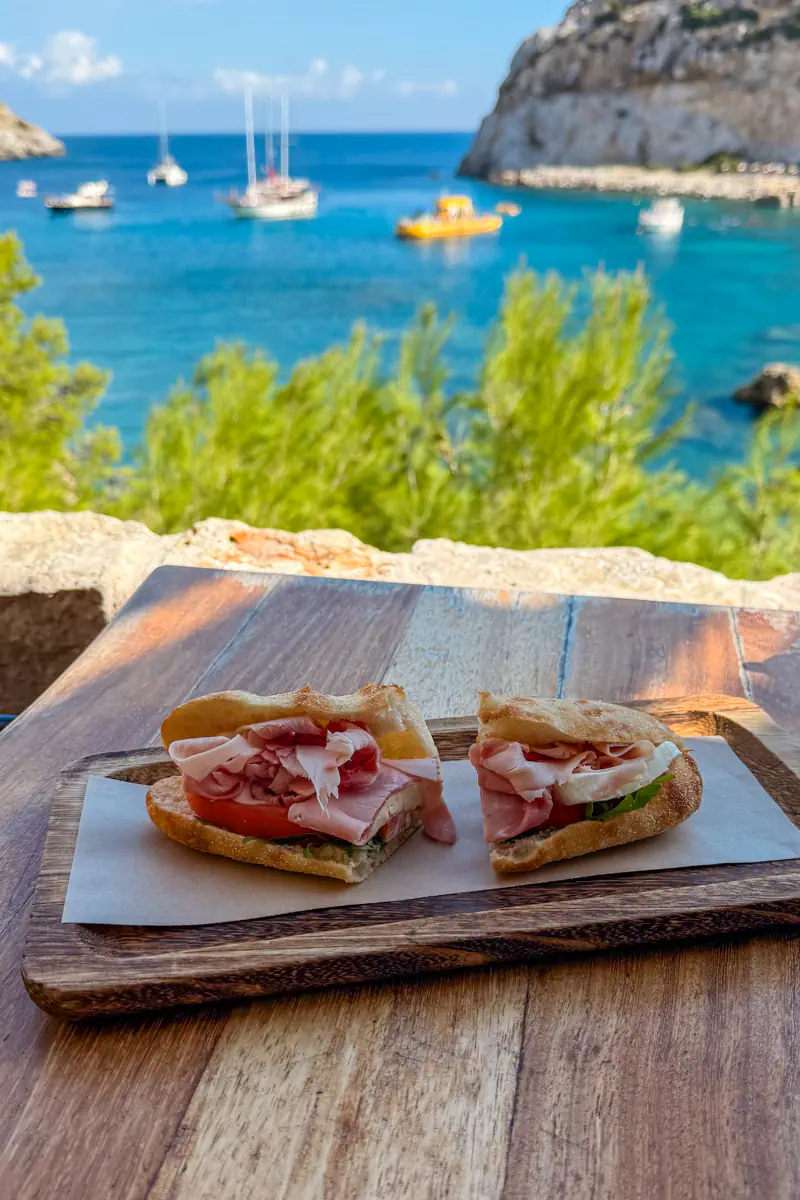 A rustic ham sandwich with tomato and lettuce on fresh bread is served on a wooden board with a backdrop of the Aegean Sea and anchored boats in the bay.