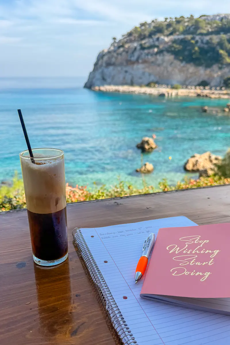 A tall glass of freddo espresso sits beside a pink notebook that says “Stop Wishing Start Doing” and a spiral-bound notepad on a wooden table overlooking the turquoise sea and rocky coastline in Rhodes.