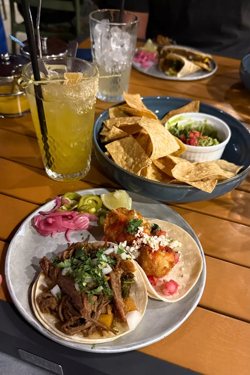 A plate of tacos with pickled red onions, meat, and cheese bites beside tortilla chips, guacamole, and a margarita at an outdoor table in Revelstoke.