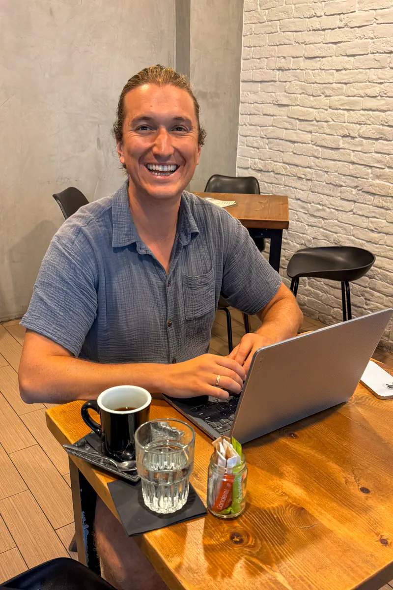 A cheerful man sits at a wooden café table working on his laptop with a black coffee, a glass of water, and sugar packets in a small jar, in a minimalist coffee shop with white brick walls.