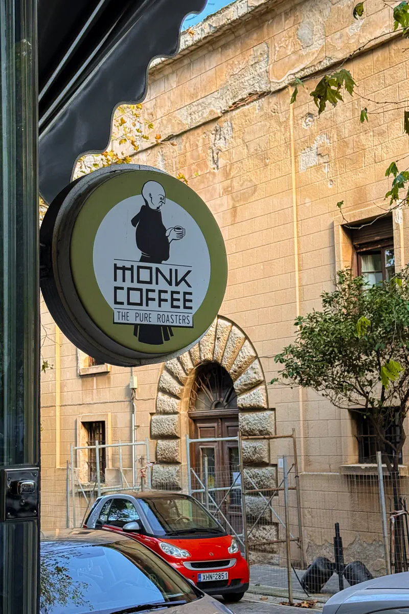 Round sign for “Monk Coffee – The Pure Roasters” hangs outside a café in Rhodes, with an old stone building, a red Smart car, and trees in the background.
