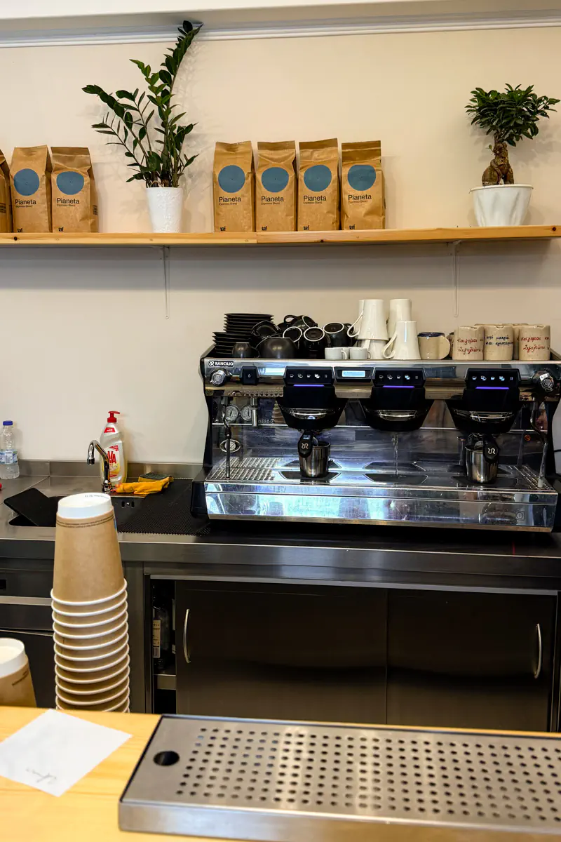 A commercial espresso machine setup inside a café in Symi, surrounded by coffee cups, paper takeaway cups, and bags of Planeta coffee beans on a shelf above.