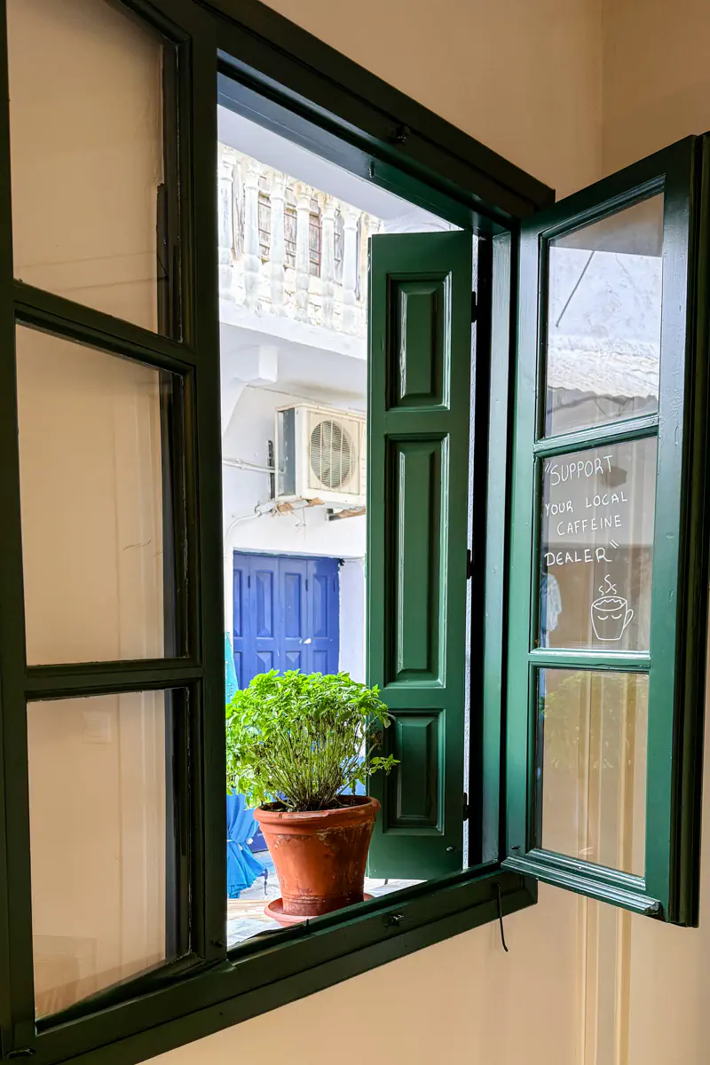 An open green window in a café reveals a potted herb plant on the sill and a blue door across the way, with a handwritten message on the glass that reads “Support your local caffeine dealer.”
