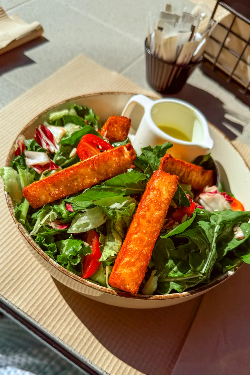A fresh mixed greens salad topped with golden grilled halloumi strips and cherry tomatoes, served with a small white jug of dressing on the side.