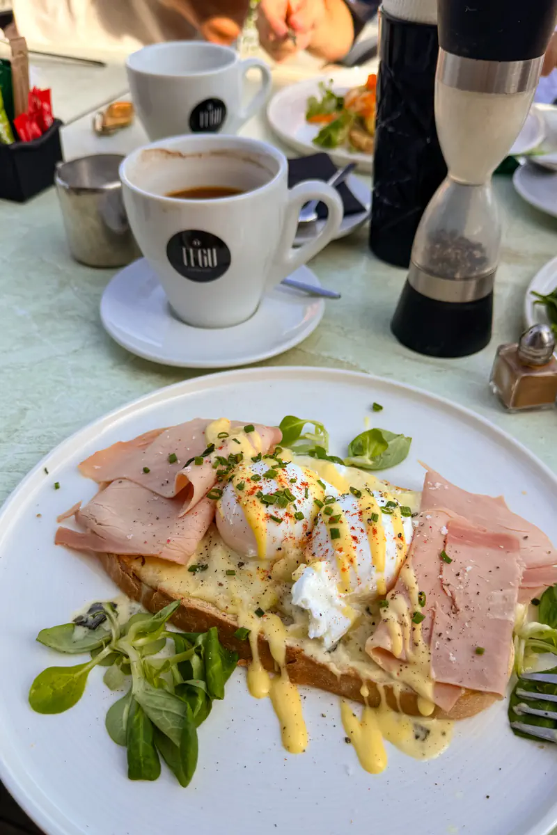 A plate of eggs benedict on toast with slices of ham, melted cheese, hollandaise sauce, and fresh greens, served alongside coffee at a café table in Rhodes.