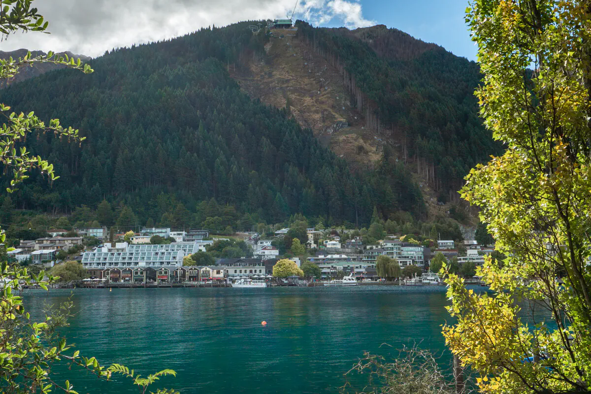 A view across Lake Wakatipu in Queenstown, with the town's waterfront buildings in the foreground and a forested mountain slope behind. The Skyline Queenstown gondola terminal is visible at the top of the hill.