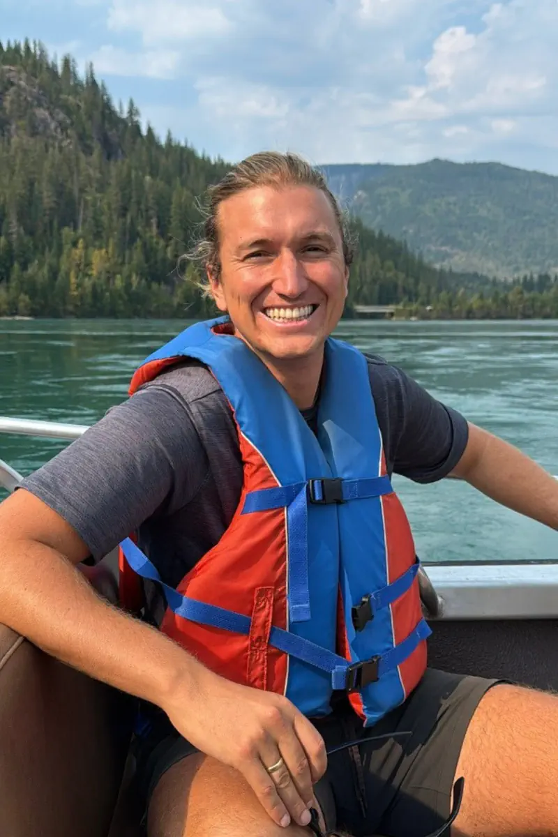 Man wearing a blue and orange life jacket smiling while sitting on a boat on a calm river, with forested mountains in the background.