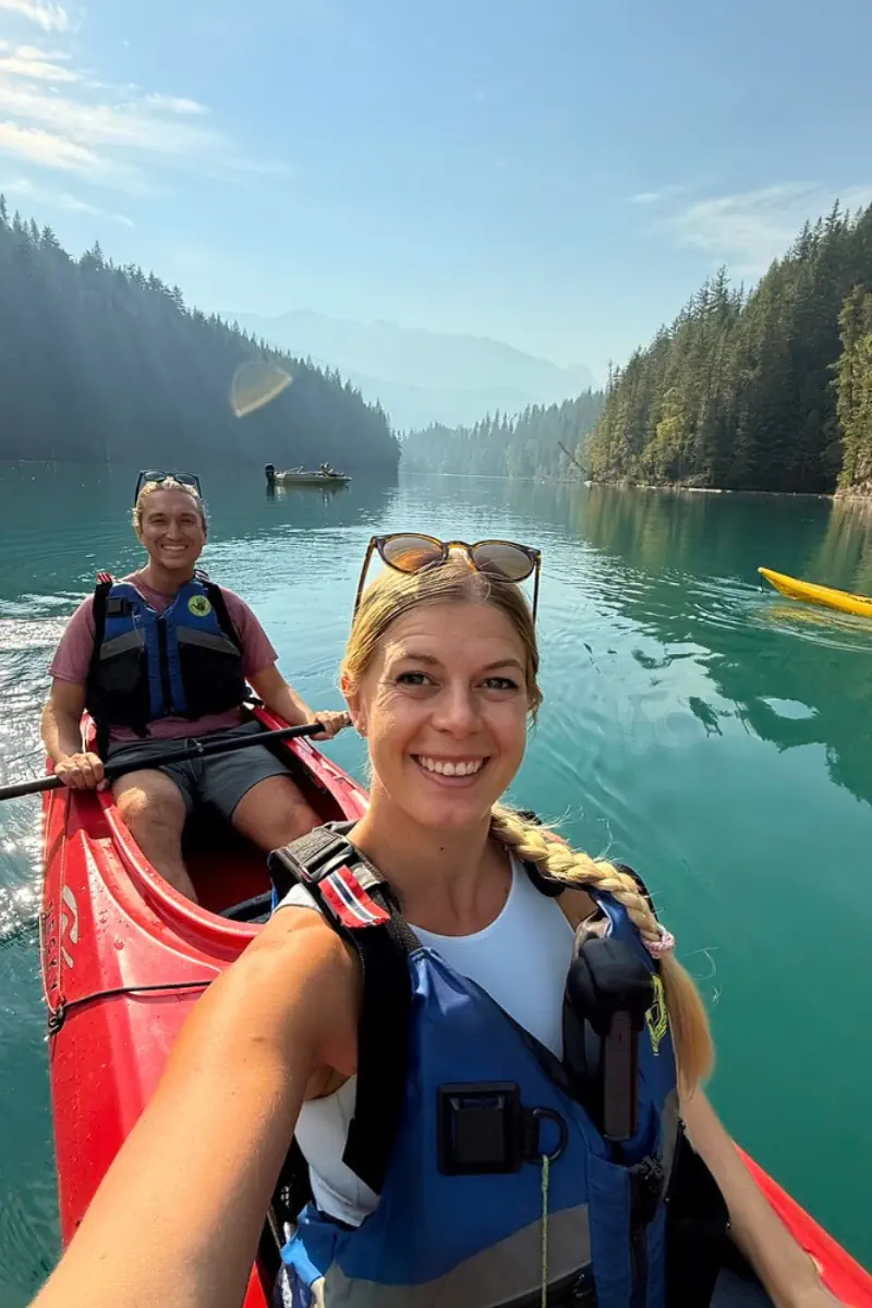 Two people kayaking on a turquoise river surrounded by forested mountains, with the woman taking a selfie from the front of the red kayak while both wear life jackets.