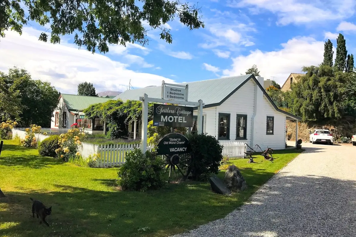 A sunny view of Settlers Cottage Motel in Arrowtown, New Zealand, showcasing white heritage-style buildings with green roofs surrounded by manicured gardens and a white picket fence.