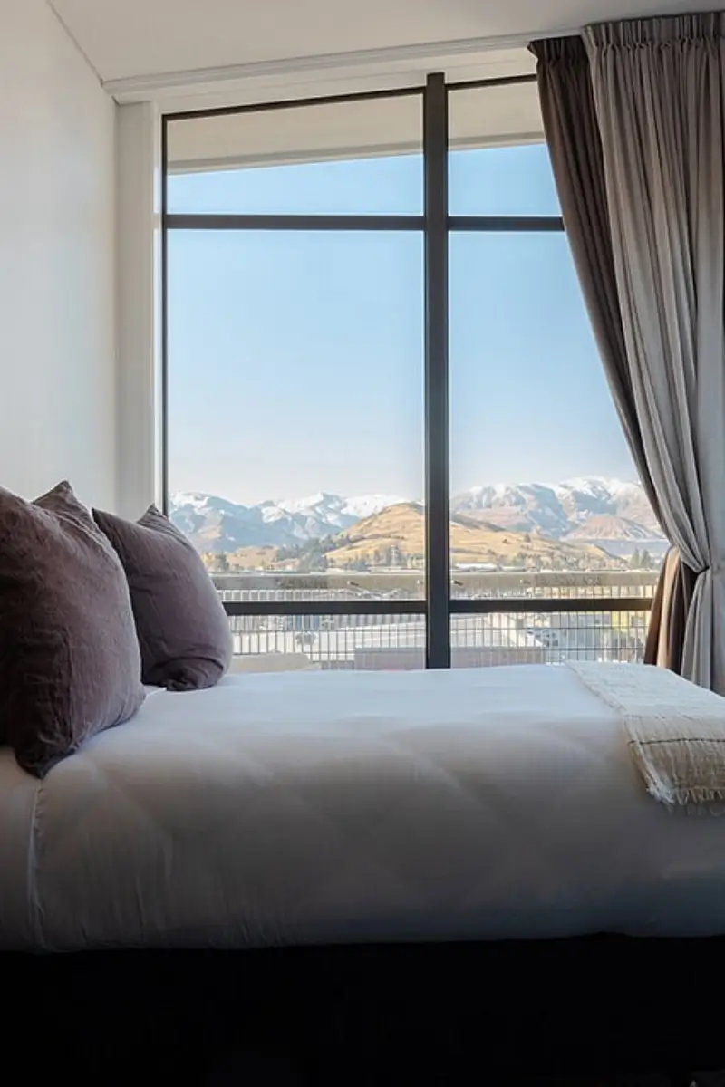 A stylish hotel room at Sudima Queenstown with a neatly made bed and dark throw pillows, featuring a large window that frames a stunning view of snow-capped mountains and rolling hills under a clear blue sky.