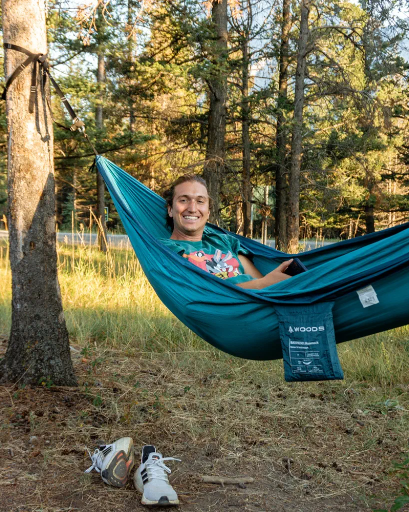 A person relaxing in a teal hammock tied between two trees in a forested area, smiling while holding a phone. The ground below is covered in dry grass and pinecones, with a pair of white running shoes placed beside the hammock. Sunlight filters through the trees, creating a warm, peaceful outdoor setting.