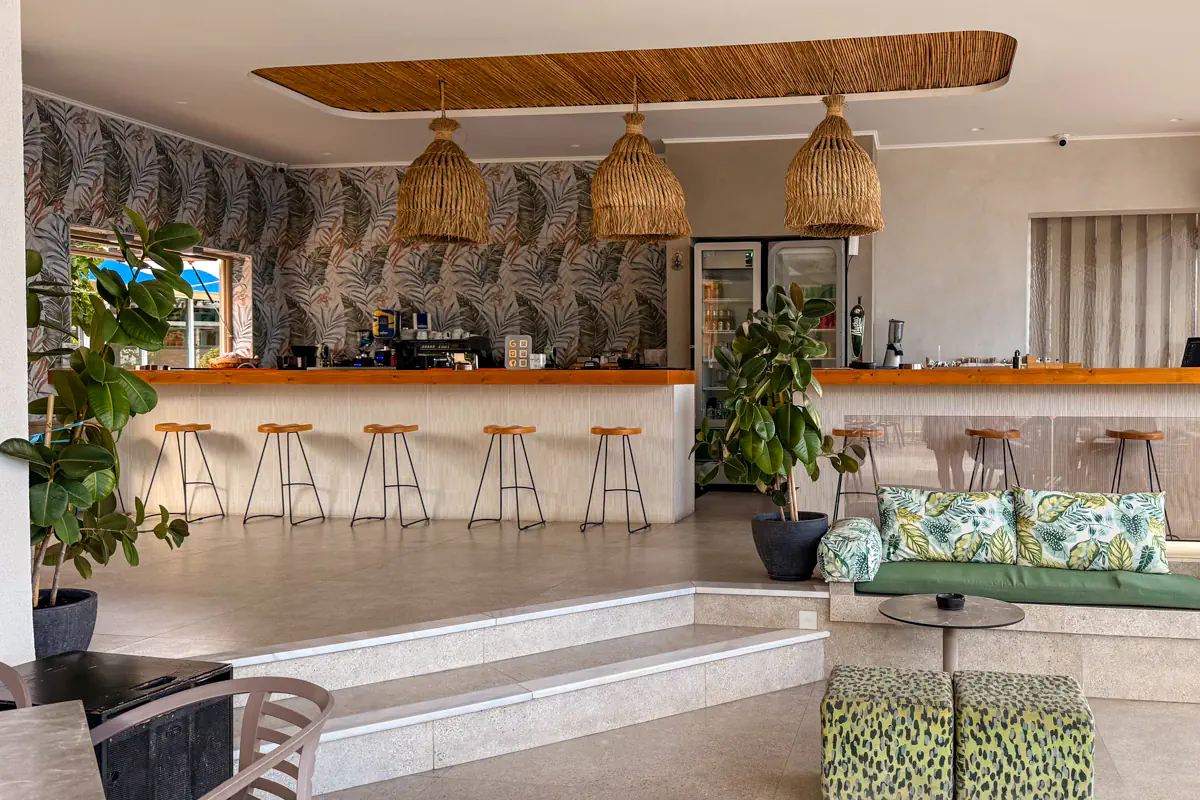 A modern open-air café interior in Rhodes featuring a long bar with wooden stools, tropical leaf wallpaper, woven pendant lights, and green plant accents with patterned seating.