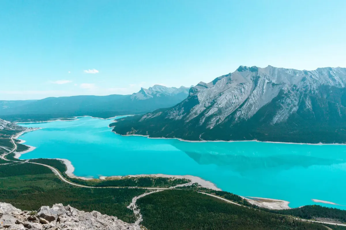 A panoramic view of a bright turquoise lake surrounded by forested valleys and dramatic grey mountains under a clear blue sky. A winding road traces the shoreline, and the jagged peaks reflect softly on the calm water.