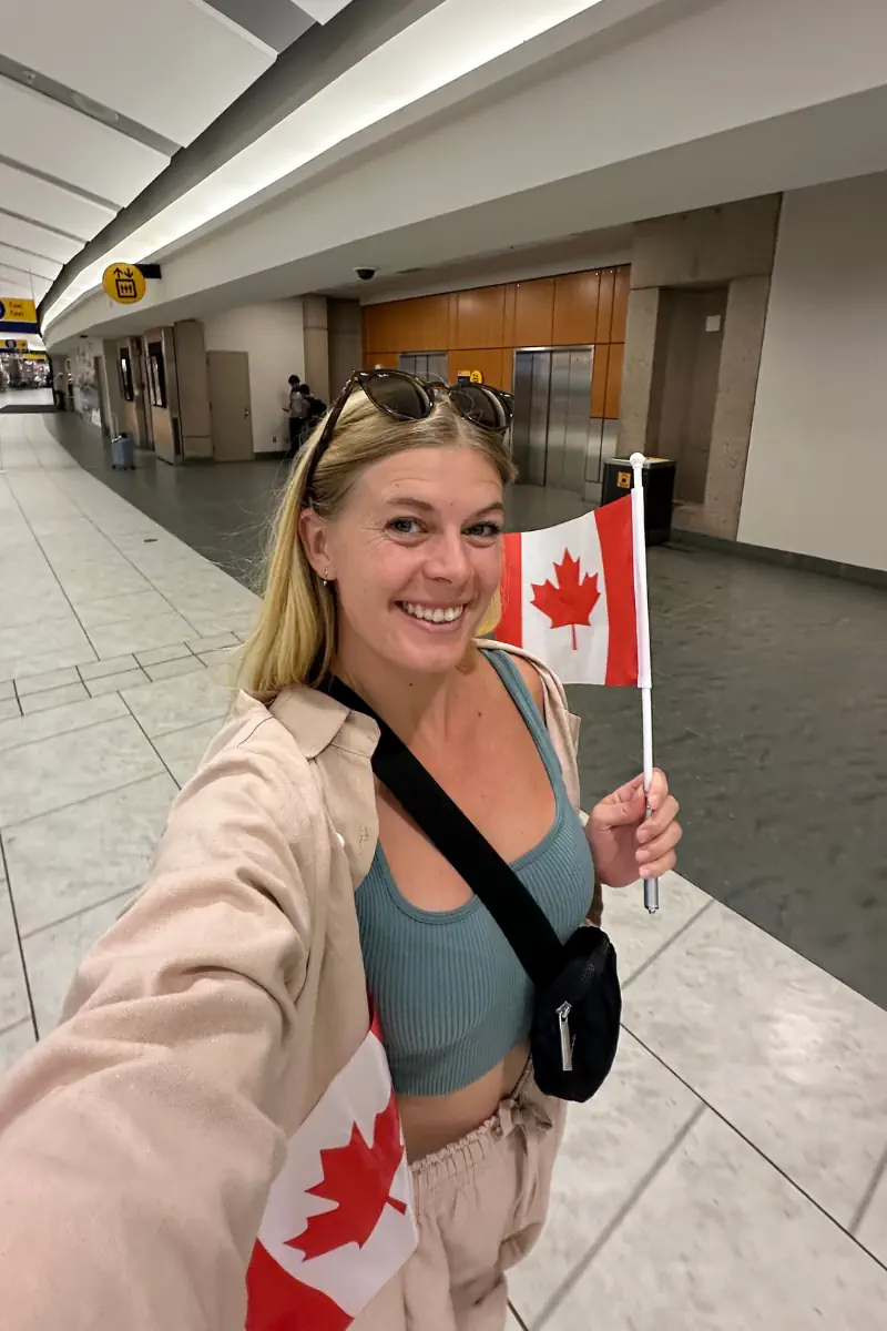 A woman standing in an airport hallway, smiling while holding a small Canadian flag. She is wearing a light-coloured outfit with a teal top, sunglasses on their head, and a black crossbody bag. The airport corridor behind them is bright and mostly empty.