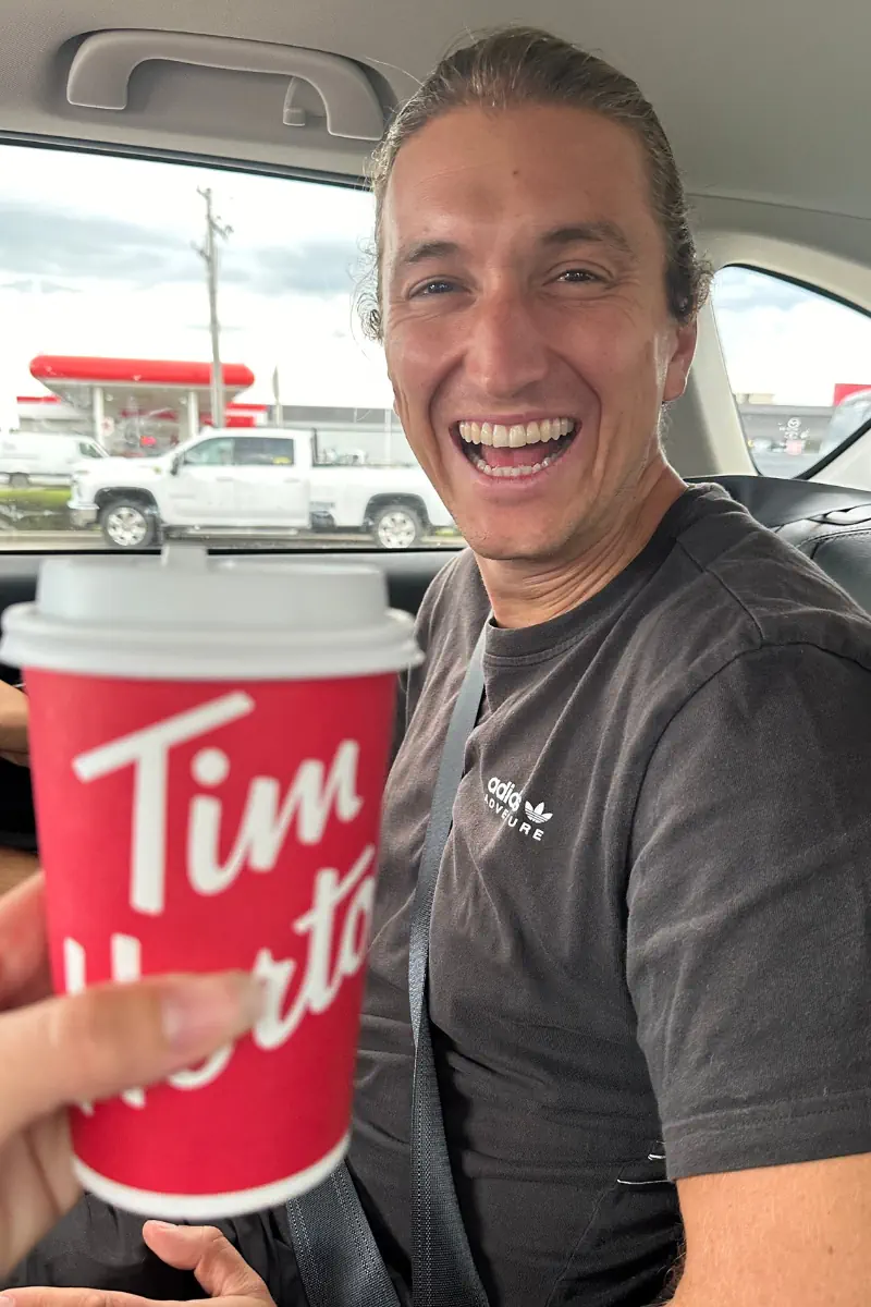 A man sitting in the seat of a car, smiling widely. In the foreground, a hand holds a red Tim Hortons coffee cup toward the camera. Through the window, a gas station and parked vehicles are visible outside.