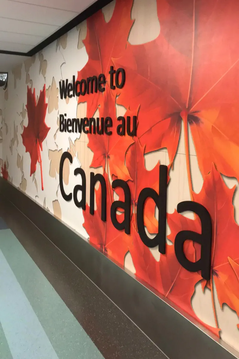 A wall inside an airport decorated with large red maple leaves and bold black lettering that reads “Welcome to Bienvenue au Canada.” The colourful design lines a hallway floor with green and blue accents.