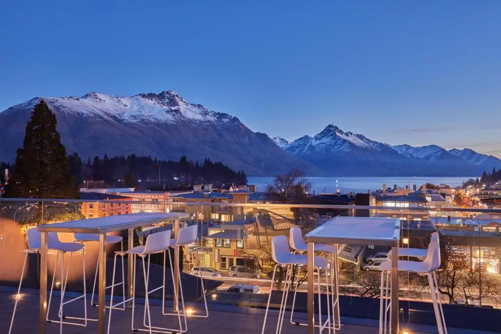 Outdoor rooftop seating with modern white tables and chairs overlooks Queenstown, New Zealand at dusk. The view includes a lit-up town, Lake Wakatipu, and snow-capped mountains under a clear twilight sky.